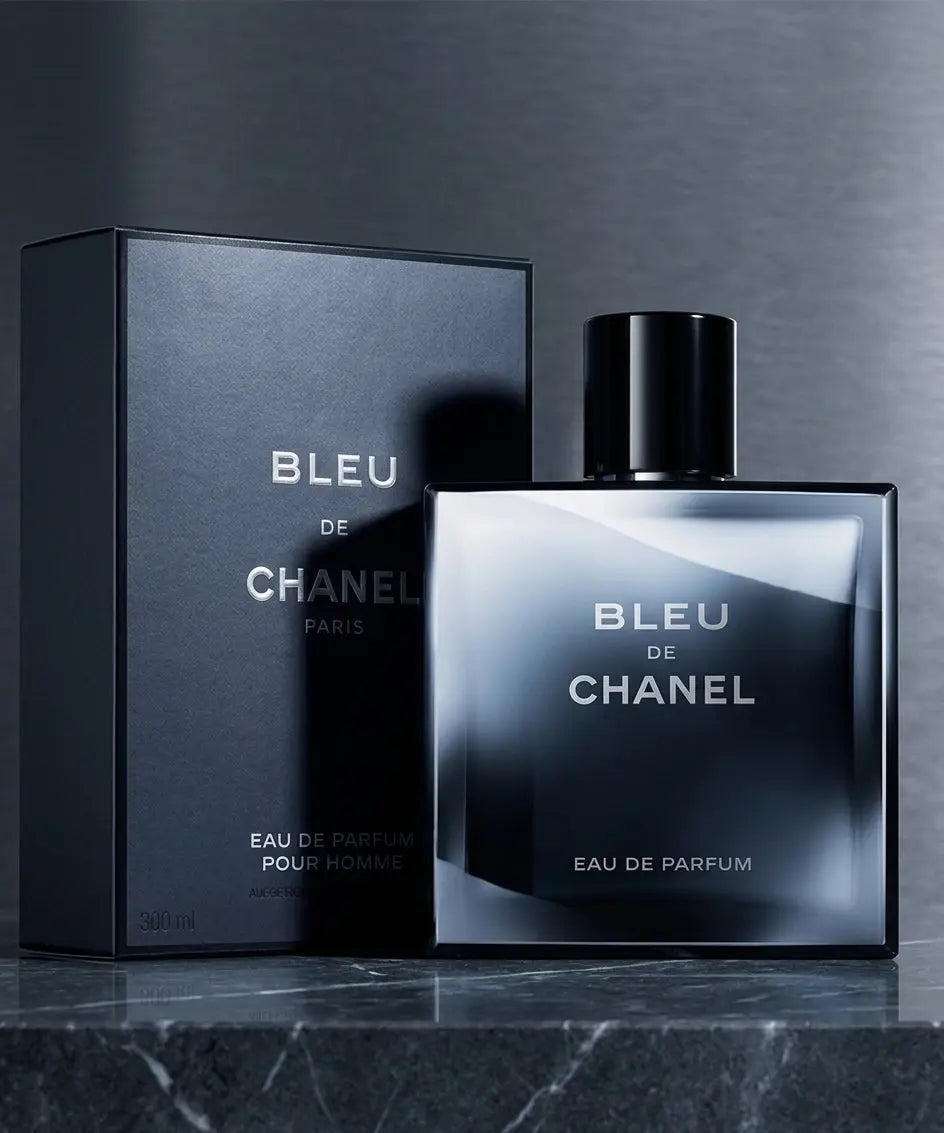 A square glass bottle of Bleu de Chanel Eau de Parfum next to its dark matte retail box on a reflective metallic surface